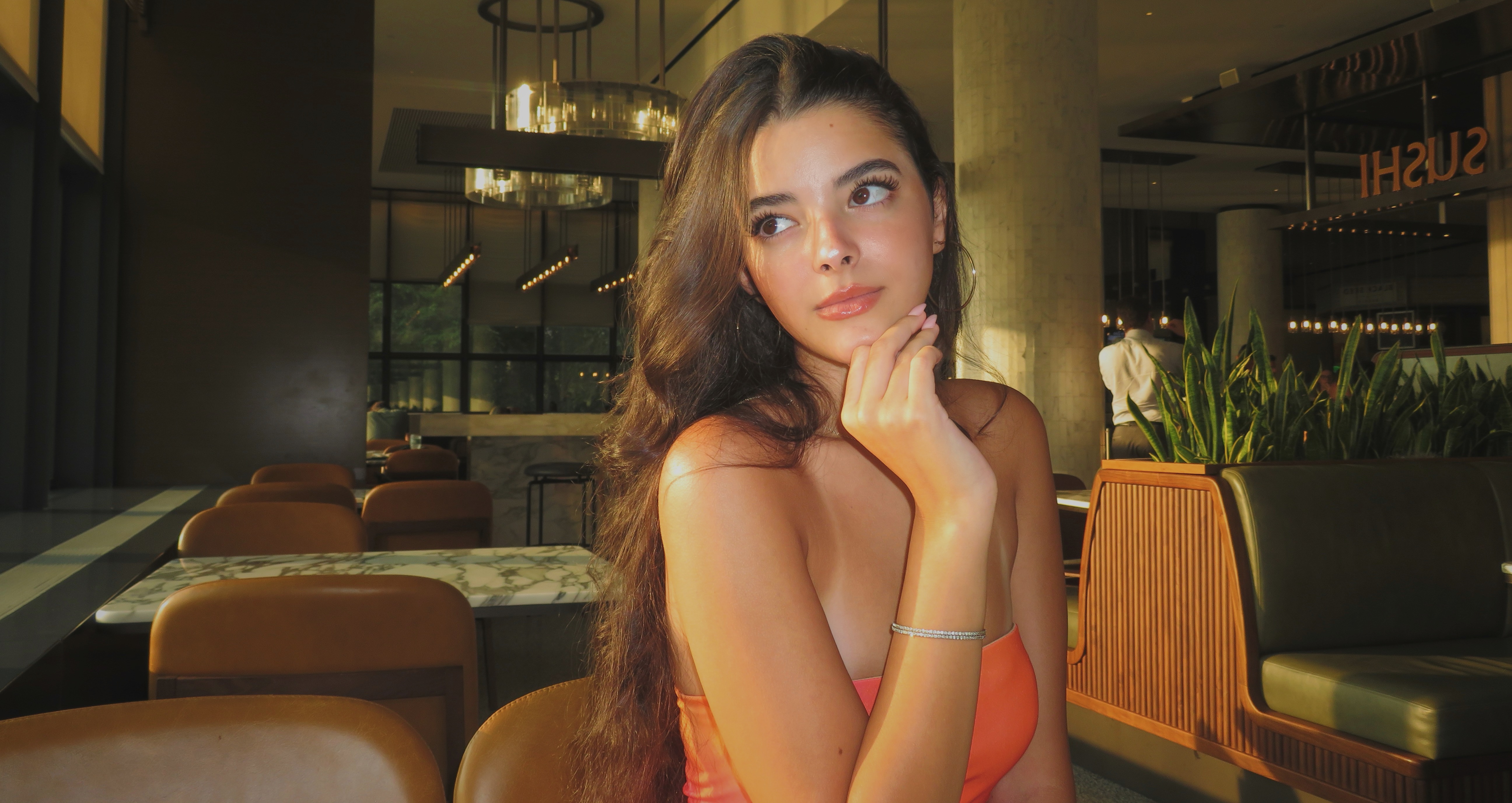 Female woman wearing an orange dress sitting down with hand to chin looking away. Brown hair parted to the side.